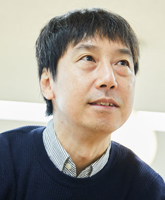A person wearing a navy sweater over a striped collared shirt, sitting indoors with soft lighting.
