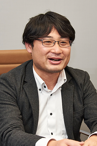 A business professional with dark hair and glasses is sitting and smiling during an interview, wearing a dark jacket and white shirt.