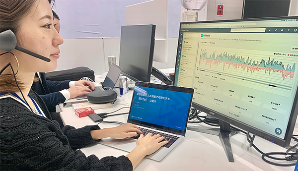 A woman with a headset works at a desk with a laptop and large monitor displaying analytics charts and data, representing user analytics in a case study for RevComm on AWS.