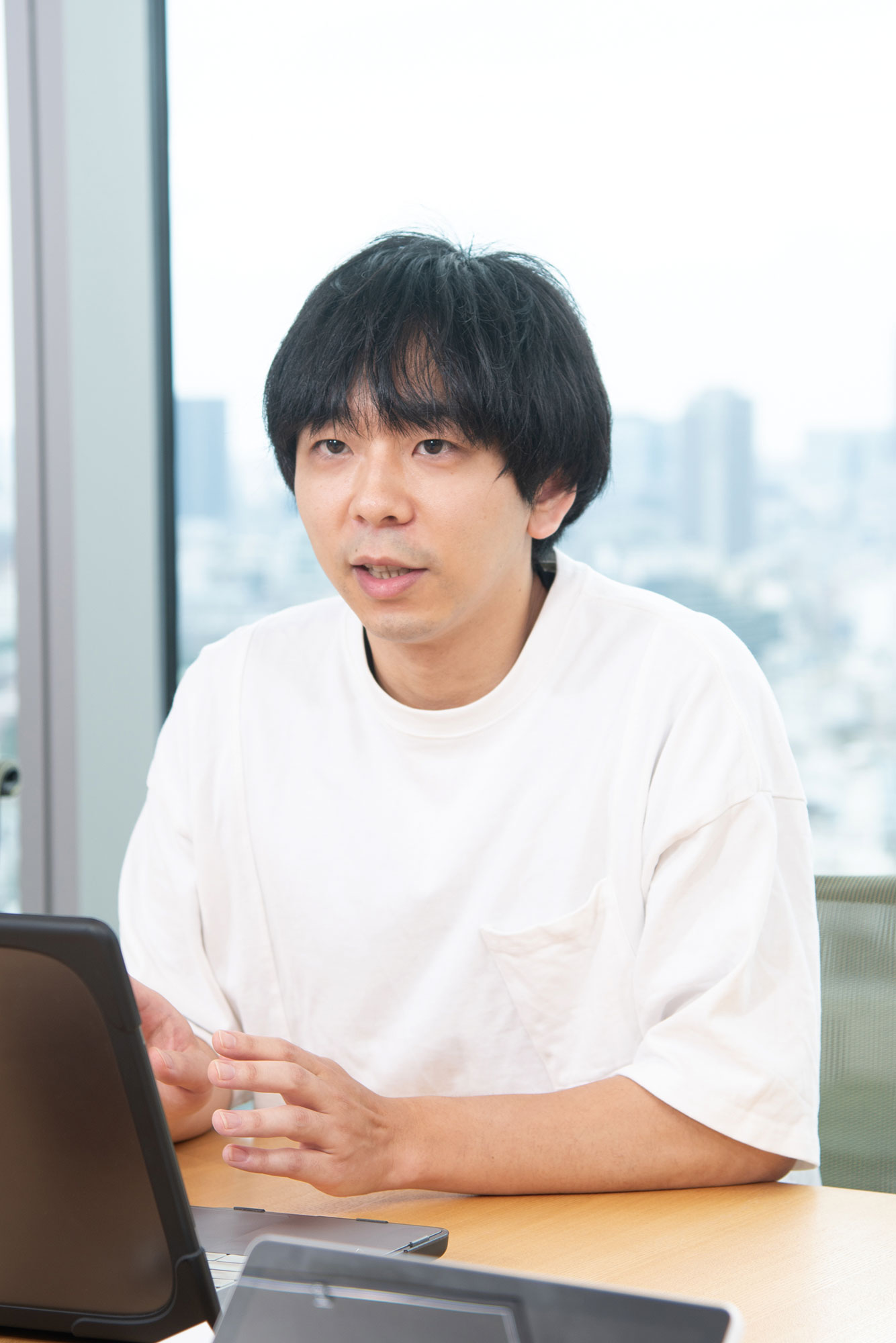 Person wearing a white shirt sitting at a desk with a laptop, in an office setting with a cityscape visible through the window.