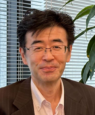 Portrait of Sato Yasuki from Toda Kensetsu, featured in an AWS solution case study. The individual is wearing glasses and a business suit, seated indoors with a plant and window blinds in the background.