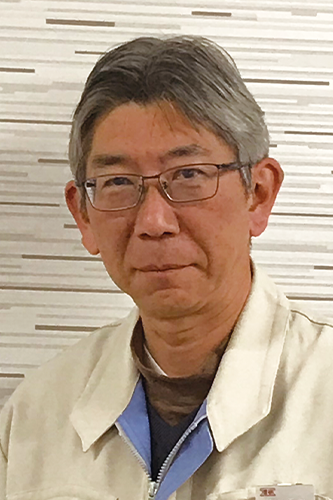 A man with gray hair and glasses wearing a beige work jacket and blue-collared shirt, standing in front of a patterned wall.