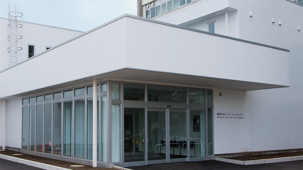 The exterior view of the Hirosaki Innovation Center for Health Promotion, featuring modern white architecture with large glass windows and an entrance area.
