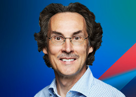 A man with curly hair and glasses is smiling in front of a blue background with red and blue geometric accents. He is wearing a light-colored patterned shirt.