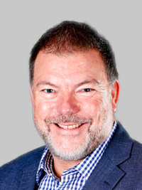 Portrait of a smiling man wearing a checked shirt and dark blazer, photographed against a plain background.