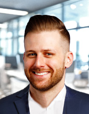 A professional headshot of a smiling man in a suit, with short hair and a beard, in a modern office environment with large windows in the background.