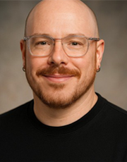 A portrait photograph of a smiling person wearing clear glasses, a beard, and a black shirt, with a neutral background.