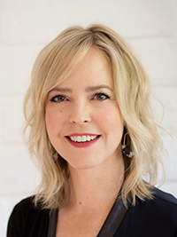 A professional headshot portrait of a smiling woman with blonde hair, wearing a black top, in front of a light background.