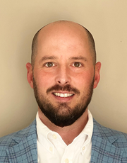 A professional headshot of a man with a beard and shaved head, wearing a blue plaid jacket and white shirt, posed against a neutral background.
