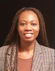 A professional woman with long braids, wearing a plaid blazer, smiling in front of a dark background.