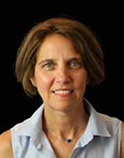 Professional headshot of a woman with short brown hair, wearing a sleeveless light blue collared top, posed in front of a plain dark background.