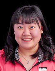 Professional headshot of a woman with black hair, wearing a pink jacket and a necklace, smiling at the camera in front of a black background.