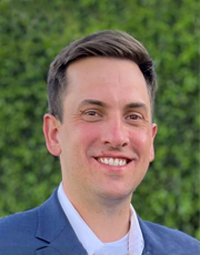 Professional portrait of a smiling man wearing a blue suit and white shirt, standing outside with a green leafy background.